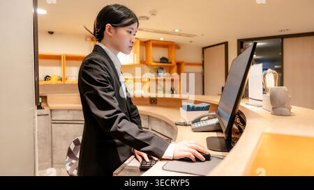 Réceptionniste professionnel taïwanais en uniforme travaille à la réception moderne, en utilisant un ordinateur et un téléphone dans un hall propre, prêt à accueillir les clients Banque D'Images