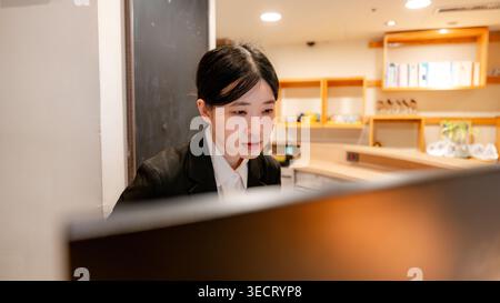 Réceptionniste taïwanais en uniforme à la réception, concentré sur l'ordinateur dans un hall moderne avec éclairage chaleureux et étagères. Banque D'Images