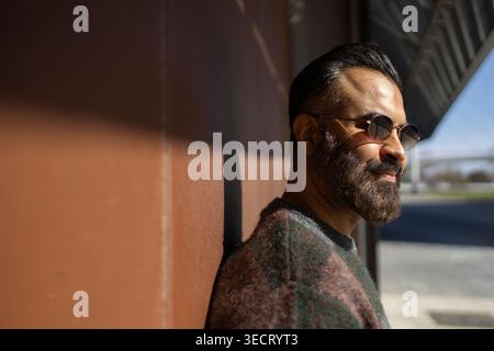 Profil latéral d'un homme arabe avec une barbe et des lunettes de soleil appuyées contre un mur à l'extérieur, lumière chaude du soleil et ombres douces créant une ville calme et moderne Banque D'Images