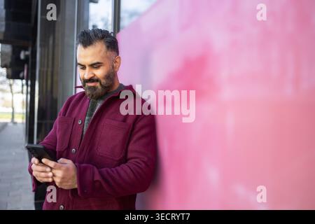 Homme arabe avec la barbe se penche sur un mur de bâtiment moderne tout en envoyant des SMS sur un smartphone à l'extérieur, portant une veste Bordeaux, avec un espace de copie pour la technologie Banque D'Images