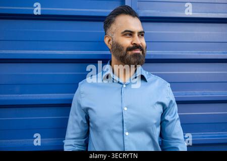 Homme d'affaires arabe confiant dans une chemise bleu clair se tient contre un mur bleu, souriant et regardant de côté. Banque D'Images