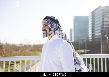 Homme d'affaires de cheikh arabe confiant en thobe blanc et keffiyeh se tient à l'extérieur par une ligne d'horizon de la ville moderne, regardant vers l'avant en plein jour, symbolisant Banque D'Images