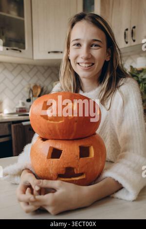 Fille souriante à l'intérieur tenant des citrouilles d'Halloween sculptées empilées sur une table de cuisine, atmosphère chaleureuse à la maison avec décor d'automne, visages jack-o'-lanterne et F. Banque D'Images