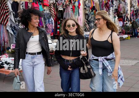 Trois joyeuses femmes diverses, deux bras de liaison, marchant et souriant devant des stands de marchandises colorées à un marché en plein air, appréciant l'amitié et le sho Banque D'Images