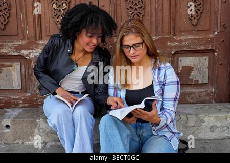 Deux amies diverses sont assises sur les marches extérieures, partageant un livre et discutant du contenu, en se concentrant sur l'éducation et l'apprentissage collaboratif près d'un Banque D'Images