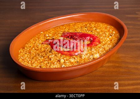 Ragoût de riz espagnol traditionnel avec un riche bouillon servi dans un plat rustique en argile, garni de crevettes rouges. Photographie de nourriture en gros plan sur une table en bois, chaud à Banque D'Images