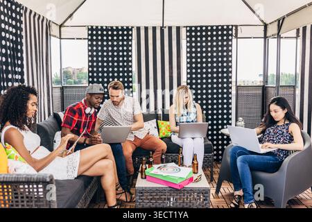 Groupe diversifié de collègues travaillant à distance sur un salon de terrasse extérieur, en utilisant des ordinateurs portables et des téléphones autour d'une table avec des boissons, collaborant dans un casu Banque D'Images