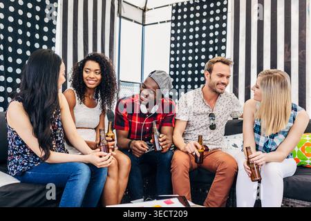Groupe diversifié de jeunes amis se relaxant sur un canapé, bavardant et riant tout en tenant des bouteilles de bière dans un salon confortable avec un décor à rayures et pois Banque D'Images