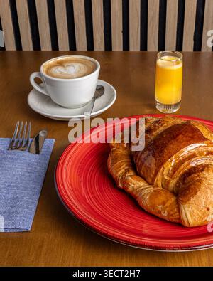 Petit déjeuner matinal confortable sur une table en bois avec un croissant au beurre sur une assiette rouge, un cappuccino avec latte art, et un verre de jus d'orange, prêt à e Banque D'Images