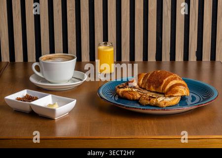 Petit déjeuner le matin sur une table en bois avec un croissant, café chaud dans une tasse blanche, jus d'orange, et de petits bols de confiture et beurre dans un café s confortable Banque D'Images
