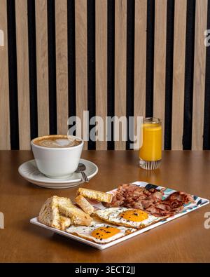 Petit-déjeuner matinal sur une table avec un oeuf frit avec pain grillé, bacon croustillant et jambon, un cappuccino dans une tasse blanche, et un verre de jus d'orange dans un coz Banque D'Images