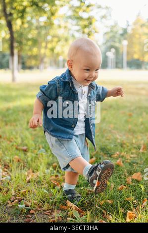 Un jeune garçon souriant court à travers l'herbe verte dans un parc ensoleillé, portant une chemise en Jean et un short. Enfant ludique appréciant l'activité de plein air, l'air frais, et Banque D'Images