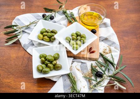 Trois bols blancs d'olives vertes à côté d'un verre d'huile d'olive sur une planche de bois, stylisé avec du romarin, du thym, des branches d'olivier et un tissu rayé sur un Banque D'Images