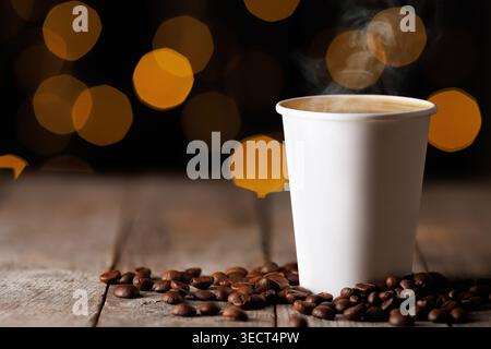 Vapeur café chaud dans une tasse de papier blanc, entouré de grains de café torréfiés sur une table en bois rustique avec des lumières bokeh chaudes Banque D'Images