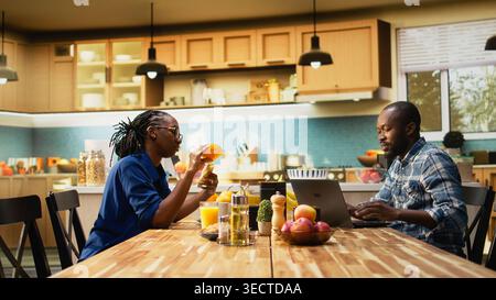 Homme noir souriant travaillant à distance à la table et discutant avec sa petite amie, buvant du café pendant que le petit ami écrit un rapport sur son ordinateur. Télétravail à la table du petit déjeuner. Banque D'Images