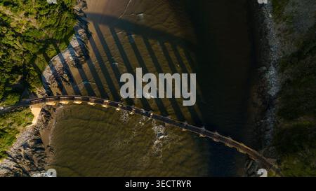 Prise de vue par drone aérien d'un pont ferroviaire historique enjambant la gorge de la rivière Kaaimans dans la nature sauvage de Garden route, avec des parois rocheuses couvertes de mousse, flowi Banque D'Images