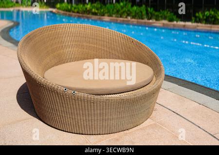 Chaise longue ronde en osier avec un siège rembourré positionné à côté d'une piscine, avec de l'eau bleu clair et une végétation luxuriante en arrière-plan Banque D'Images