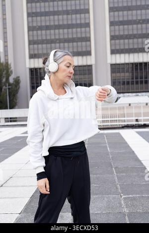 Femme mature debout sur la terrasse sur le toit vérifiant le dispositif de poignet, portant des écouteurs et un sweat à capuche blanc Banque D'Images
