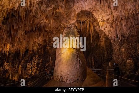 Une majestueuse stalagmite s'élève au centre d'une caverne, illuminée par une lumière dorée tandis que les visiteurs marchent le long des balustrades métalliques environnantes. Banque D'Images