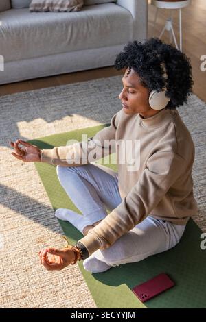 Femme afro-américaine dans ses années 30 assise les jambes croisées sur un tapis de yoga vert méditant avec des écouteurs Banque D'Images