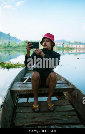 Un jeune homme portant un chapeau seau rose et une veste sombre est assis dans un bateau traditionnel en bois sur un lac calme, capturant une photo avec son smartphone. Banque D'Images
