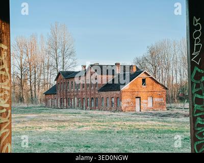 Riga, Lettonie - 30 avril 2026 : ancien bâtiment industriel en briques rouges abandonné dans un champ rural calme, marqué de graffitis et de pourriture. Atmospheri Banque D'Images