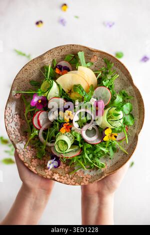 Délicieuse salade d'été avec des fleurs comestibles, des légumes, des fruits, des microverts et du fromage. Femme tenant assiette avec salade, vue de dessus. Manger propre et sain Banque D'Images