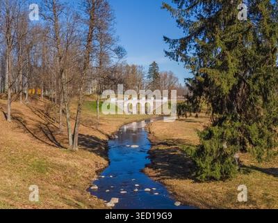 Magnifique parc de printemps. La lumière du soleil jaillit de l'eau calme de la rivière. Les arbres sont hauts sur les deux rives. Le pont vole gracieusement à travers le ruisseau. Le ciel bleu s'étend au-dessus de la scène. Les feuilles d'automne couvrent le sol. Oranienbaum Banque D'Images