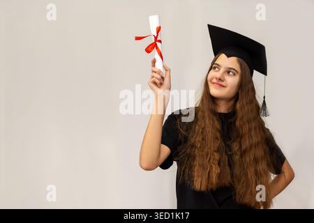 Heureux diplômé tenant le diplôme vers le haut. Jeune femme souriante dans la casquette de graduation élève un diplôme roulé avec ruban rouge, célébrant la réussite scolaire et Banque D'Images