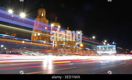 Une photo nocturne de la cathédrale de Varna en exposition longue montre des dômes et une façade illuminés avec des traînées de lumière provenant de voitures qui passent, créant des lignes dynamiques dans la for Banque D'Images