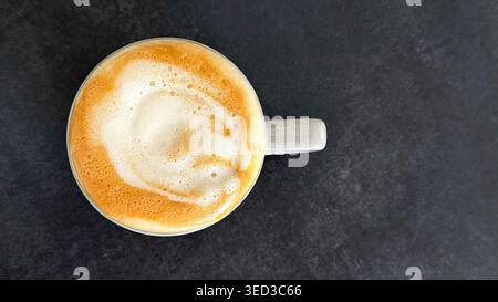 Vue rapprochée d'une tasse à café en céramique blanche remplie de cappuccino mousseux sur une surface texturée sombre, présentant un design artistique latte complexe sur le dessus Banque D'Images