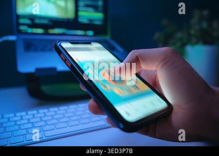 Gros plan de la main tenant un smartphone et faisant défiler rapidement le flux de médias sociaux dans une pièce sombre avec un bureau sur fond, éclairé par une lumière bleue. Dépendance numérique et consommation de contenu sans esprit Banque D'Images