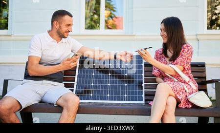 Homme et femme assis sur le banc, souriant et utilisant des smartphones, connectés à un panneau solaire. Concept d'énergie renouvelable durable, démontrant l'utilisation pratique de l'énergie solaire pour les dispositifs de charge. Banque D'Images
