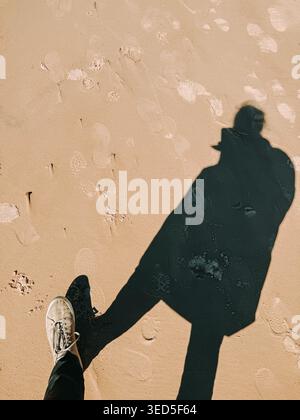 Vue de dessus créative de l’ombre et de la sneaker d’une personne sur une plage de sable avec des empreintes visibles. La forte lumière du soleil crée un contraste audacieux, idéal pour les concepts o Banque D'Images