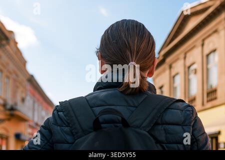Voyageur seul avec sac à dos et queue de cheval marche dans une rue historique de la ville vue de derrière. Évoque l'exploration urbaine, les trajets quotidiens, la planification de voyages et Banque D'Images
