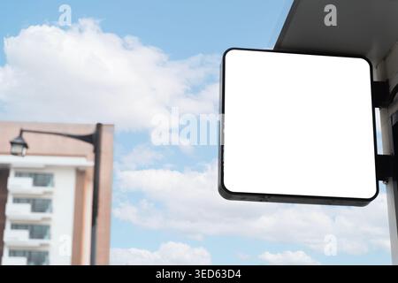 Panneau blanc carré de devanture attaché à l'extérieur d'un bâtiment avec ciel bleu et fond urbain doux, idéal pour la marque de détail, affichage de logo, adverti Banque D'Images