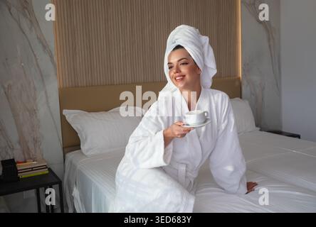 Belle femme assise sur le lit tenant le café. Portrait doux avec serviette sur la tête. Ambiance matinale calme dans la chambre d'hôtel. Concept de style de vie relaxant du spa. Peau Banque D'Images