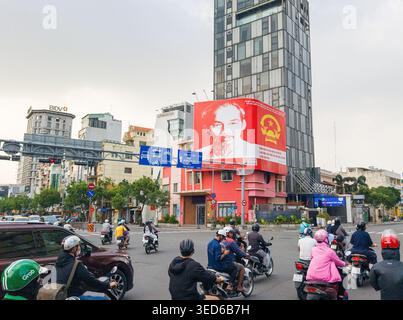 Intersection animée à Ho Chi Minh-ville, caractérisée par un trafic dense de motos et des bâtiments modernes de grande hauteur.Un grand panneau d'affichage de propagande rouge représentant l'image de Ho Chi Minh est bien visible sur l'un des bâtiments. Ho Chi Minh ville, Vietnam, Asie Banque D'Images