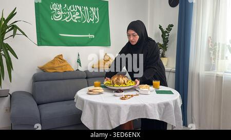 Une femme dans une abaya noire et un hijab place un poulet rôti sur une table pour une célébration. Dans un salon lumineux, un drapeau saoudien est suspendu en évidence, Banque D'Images