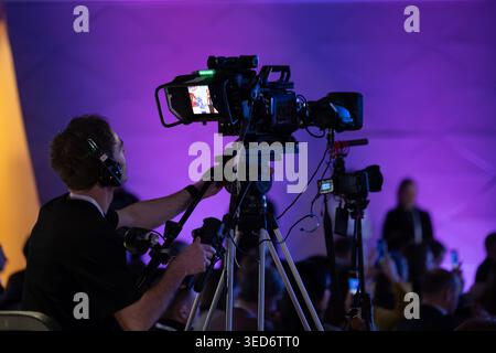 Cameraman opérant caméra vidéo professionnelle sur trépied à l'événement en direct, éclairage de scène violet, public en arrière-plan. Banque D'Images