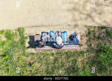 Vue aérienne de haut en bas d'un homme âgé aux cheveux gris assis sur un banc de parc et manœuvrant un drone avec un contrôleur de drone portatif. Printemps ensoleillé. Copier Banque D'Images
