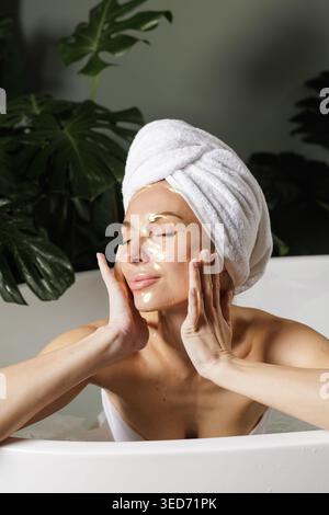 Une femme se détend dans une salle de bain moderne tout en appliquant un masque facial en or, incarnant l'auto-soin et le rajeunissement dans un environnement serein. Banque D'Images