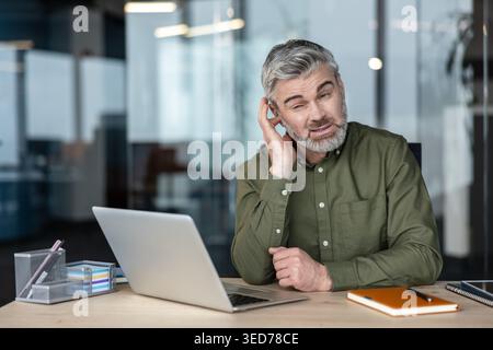 Homme d'affaires d'âge moyen paraissant confus et mettant la main à l'oreille, ayant des problèmes auditifs ou ayant du mal à comprendre l'information tout en travaillant sur un ordinateur portable dans un bureau moderne Banque D'Images