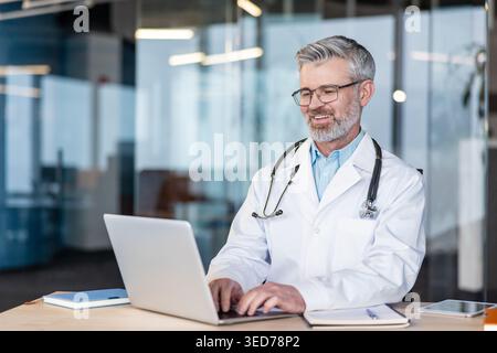Médecin en manteau blanc et stéthoscope souriant tout en tapant sur le clavier de l'ordinateur portable, en fournissant une consultation médicale en ligne ou en gérant les dossiers des patients dans un cadre de bureau moderne Banque D'Images