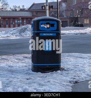 Ypsilanti, États-Unis - 12 février 2026 : bac de recyclage public sur le trottoir enneigé par la rue de la ville, scène urbaine hivernale avec des bâtiments en briques et une croix piétonne Banque D'Images