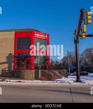 Ypsilanti, États-Unis - 12 février 2026 : bâtiment moderne du musée de pompiers en verre rouge sur un coin de rue enneigée sous un ciel bleu clair, avec des feux de circulation Banque D'Images