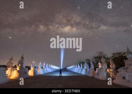Vue arrière silhouette d'un homme tenant une torche debout au milieu d'un chemin bordé de statues blanches de Bouddha la nuit, Wat Phra That Choeng Chum Worawihan, Sakon Nakhon, Thaïlande Banque D'Images
