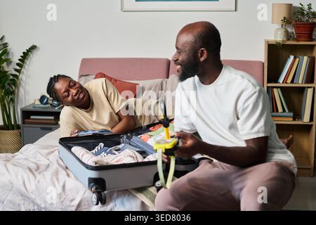 Couple noir emballant la valise sur le lit, se préparant pour le voyage et vérifiant les articles de voyage à la maison. Concept de planification de voyage, idéal pour la réservation de vacances, le tourisme, le marketing de relocalisation Banque D'Images
