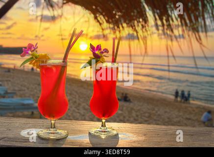 Cocktails tropicaux rouges dans un bar de plage au coucher du soleil avec des décorations de fleurs et de paille Banque D'Images