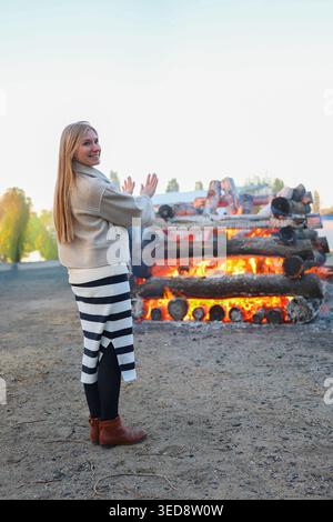 Touriste féminine ou résidente locale apprécie le feu de joie traditionnel en plein air, souriant et réchauffant les mains dans les flammes vives du soir. Banque D'Images